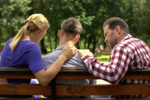 parents comforting teen while sitting on park bench on sunny day during crisis intervention